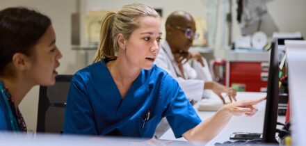 nurse-pointing-at-computer-screen-while-discussing-patient-care-with-colleague-medical-staff-working-together-in-busy-hospital-setting-healthcare-professionals-focused-on-providing-best-treatment